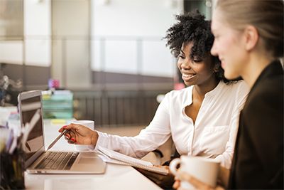 African american professional woman smiles and explains information on laptop to caucasian professional woman